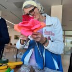 Starejša gospa z očali in belo naglavno trakico drži v rokah veliko rožo iz papirja ali blaga, ki jo natančno pregleduje in oblikuje. Oblečena je v svetlo modro in belo jopico. Pred njo na mizi ležijo barvni papirji, škarje in drugi pripomočki za ustvarjanje. Okolica je svetla in moderna, kar nakazuje na prostor skupinske delavnice.