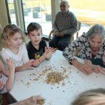V svetlem prostoru ob veliki stekleni steni je za mizo zbranih več otrok in starejših ljudi. Na mizi so drobtine in masa za oblikovanje, otroci in starejši sodelujejo pri izdelavi kroglic iz mase. Starejša gospa v črno-beli majici se sklanja k mlajšemu dečku in mu pomaga oblikovati maso.