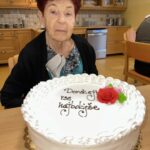 Na sliki je starejša gospa z rdečkasto barvnimi lasmi, oblečena v črn pulover in pisano bluzo, ki sedi za mizo. Pred njo je velika bela torta, okrašena z napisom "Doroteji vse najboljše" in rdečo rožo iz sladkorne mase. Gospa je resno izraza, v ozadju pa je kuhinjski prostor z lesenimi omaricami in gospodinjskimi pripomočki.
