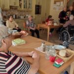 Skupina starejših moških stoji in poje v dnevnem prostoru doma za starejše. Okrog miz sedi več starejših ljudi, od katerih jih nekaj sedi na invalidskih vozičkih. V ozadju je kuhinja z osebjem in omarami.