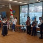 V svetli jedilnici nastopa skupina glasbenikov z diatoničnimi harmonikami in pihalom iz medenine (najverjetneje baritonom). Stojijo ob velikih oknih, oblečeni v tradicionalna oblačila, medtem ko občinstvo, večinoma starejši ljudje, sedi in uživa v nastopu. Ena gospa v folklorni obleki stoji pred skupino in nastopa z rekvizitom.