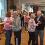 Starejši par pleše v prostoru, oba nasmejana. Moški nosi temen pleten pulover, gospa pa ima oblečeno rožasto bluzo. V ozadju nastopajo glasbeniki in še drugi pari, ki plešejo ali opazujejo dogajanje.