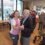 Zaposlena pleše s starejšim moškim. Oba sta nasmejana, zadaj pa se v svetlem prostoru vidi še več ljudi, ki plešejo ali opazujejo dogajanje. Na steni visi ura, pod njo pa so okraski in napisi.