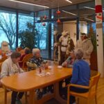V prostoru z velikimi steklenimi okni sedi skupina starejših ljudi ob leseni mizi, na kateri so skodelice in vrč z vodo. Ob mizi stojita Dedek Mraz v kožuščku z dolgo belo brado in njegov spremljevalec, oblečen v svetel plašč. V ozadju so praznične dekoracije, kot so viseče rdeče krogle in snežak na stebru.
