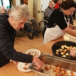 Starejša gospa v temnem puloverju z vilicami jemlje ocvrte miške iz velike kovinske posode na mizi in jih polaga na bel krožnik. Ob njej sta dve drugi gospe v predpasnikih, ena meša testo v beli plastični posodi, druga pa cvre miške v veliki črni ponvi. V ozadju so v jedilnici vidni drugi starejši ljudje, nekateri sedijo na invalidskih vozičkih.