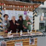 Pri stojnici stoji skupina štirih starejših gospa. Vsaka izmed njih v rokah drži izdelek s stojnice, kot so piškoti ali majhni darilni predmeti. V ozadju so informativne table, ki prikazujejo obvestila in plakate. Stojnica je okrašena z belo-črnimi trakovi in prazničnimi obeski.