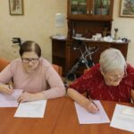 Dve starejši gospe sedita za mizo in pišeta na bele liste papirja. Ena ima oblečen rdeč vzorčast pulover, druga svetlo roza majico. V ozadju je pohištvo in več slik na steni. Na tleh so zložene hojce.