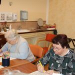 Dve starejši gospe sedita za mizo in pišeta; ena izmed njih uporablja rumeni svinčnik in barva vzorec na listu. Miza je polna barvic, papirjev in posodic. V ozadju je umivalnik in kuhinjski elementi. Na steni visijo letaki in navodila.