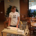 Ženska v beli majici, na kateri piše "S.P. SREČNA (penzionistka)", stoji poleg vozička s torto. Torta je pravokotne oblike, okrašena z rdečim napisom "Hvala" ter zelenimi in rdečimi dodatki. Poleg nje sta še dve ženski, ki se pripravljata na rezanje torte.