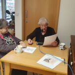 Dve starejši gospe sedita za leseno mizo v sobi z veliko dnevne svetlobe. Ena od njiju drži papir in bere v mikrofon, medtem ko druga pozorno posluša. Na mizi sta dve skodelici s toplimi napitki ter nekaj listov s besedilom. V ozadju je omara z lesenimi vrati, na kateri stoji otroška igrača, in steklena vrata, skozi katera je viden zunanji prostor s drevesi.