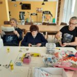 Trije starejši ljudje sedijo za mizo in se ukvarjajo z ustvarjalnimi dejavnostmi. Ena oseba bere časopis, druga lista po reviji, tretja pa se igra s pisanimi sestavljankami. Na mizi so lepilo, škarje, papirnati izrezi in škatla z dodatki. V ozadju so telovadni pripomočki in knjižne police.