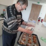 Starejši moški v puloverju s črno-belim vzorcem posipa sveže pečene kolačke s sladkorjem v prahu. Pladenj z dišečimi kolački je na beli kuhinjski površini. V ozadju je sodobna kuhinja z lesenimi vrati, lončnicami in rdečim gasilnim aparatom.