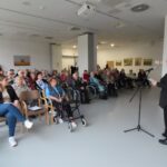 Mlada ženska z dolgimi rdečimi lasmi stoji pred mikrofonom in bere iz lista. Pred njo sedi večja skupina ljudi, starejši in mlajši udeleženci, nekateri uporabljajo pripomočke za hojo. Prostor je svetel, na stenah visijo slike, v ozadju pa je garderoba z obešalniki.
