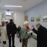 Prikazuje hodnik s slikami pokrajine, ki visijo na stenah. Skupina ljudi, večinoma starejših, si ogleduje razstavo. Nekateri sedijo na udobnih foteljih s cvetličnim vzorcem, drugi stojijo in se pogovarjajo. Na stropu visi televizijski zaslon, ki prikazuje posnetek dogodka.