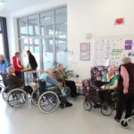 Več starejših oseb se zadržuje v svetli avli, kjer je nekaj miz in stolov. Nekateri sedijo v invalidskih vozičkih, drugi uporabljajo hodulje. Na mizi so pijače in prigrizki, v ozadju so oglasne deske s pomembnimi informacijami. V kotu stoji velika rdeča božična zvezda v lončku.