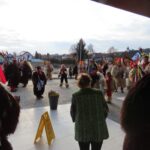 Skupina kurentov na ploščadi pred stavbo. Imajo velike maske in oprave iz ovčje volne, na glavi nosijo visoke kape s peresi in pisanimi trakovi. Nekateri držijo zvonce. V ozadju so hiše in cerkev, okoli vhoda pa cvetlični lonci.