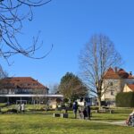 Park z več starejšimi osebami, ki se sprehajajo ali sedijo v invalidskih vozičkih. Spremljajo jih skrbniki. V ozadju je večja stavba z rdečo streho in balkoni, obdana z drevesi in zelenico. Nebo je jasno in modro.