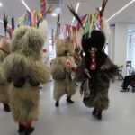 Skupina kurentov v tradicionalnih pustnih opravah pleše v krogu v svetlem prostoru doma za starejše. Oblečeni so v kožuhe, okrašene z zvonci, peresi in pisanimi trakovi. Okoli njih sedijo stanovalci doma, nekateri v invalidskih vozičkih, drugi na stolih, in opazujejo dogajanje.