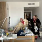 Dva moška oblečena v tradicionalni kurentovski opravi stojita ob bolniški postelji, kjer starejši moški leži in se rokuje z enim od njiju. Na obrazu ima nasmeh, ki izraža veselje ob obisku. Bolniška soba je urejena, z leseno omarico in medicinsko opremo.