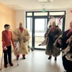Starejša gospa s pomočjo bergel stoji ob treh kurentih, ki so oblečeni v tradicionalne oprave, vsak drži v rokah leseno gorjačo in robce. Gospa se nasmehne in ponosno pozira skupaj s pustnimi liki. Za njimi so steklena vrata, ki vodijo na balkon ali teraso doma.