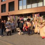 Skupinska fotografija pred stavbo: v ozadju stojijo kurenti z barvitimi trakovi na glavah, pred njimi pa stanovalci in obiskovalci, nekateri na invalidskih vozičkih.