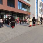 Skupina kurentov s pisano okrašenimi glavami pleše in razveseljuje ljudi, ki so zbrani pred sodobno stavbo, med njimi tudi osebe na invalidskih vozičkih.