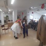 Oseba v klobuku in modrem predpasniku z ročnim košem ter oseba v belem pustnem kostumu z barvnimi trakovi hodita po dvorani, ob njima pa sedijo in stojijo starejši ljudje.