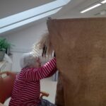 Starejša oseba v rdeči črtasti majici sedi na stolu in pomaga pri oblikovanju velikega pustnega kostuma iz vrečevine.