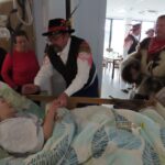 Starejša gospa leži v postelji in se nasmehne, ko ji moški v tradicionalni noši drži roko. Okoli nje stojijo še drugi obiskovalci, eden od njih je oblečen v kožuh s kravjimi zvonci. Moški v noši nosi črn klobuk s peresi in barvitimi trakovi.