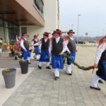 Moški v narodnih nošah zapuščajo stavbo in korakajo po tlakovanem pločniku. Držijo se za venec iz zelenja. V ozadju je sodobna zgradba z velikimi okni, na levi strani pa oseba v tradicionalni ovčji kožuhovini. Okoli vhoda so lončnice s pisanimi rožami.
