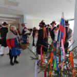 V notranjem prostoru stoji skupina moških, oblečenih v tradicionalne narodne noše. Nosijo bele srajce, črne telovnike, modre predpasnike in črne klobuke z barvitimi trakovi. V ospredju stoji ženska v rdečem plašču in modrem predpasniku, ogrnjena s pleteno ruto, ter drži košaro. Na desni strani slike je majhna smrečica, okrašena s pisanimi trakovi in slovensko zastavo.