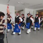 Skupina možakarjev v tradicionalnih oblačilih koraka skozi hodnik, eden od njih drži venec iz zelenja. Ob strani jim maha starejša gospa v rjavem puloverju, naslanjajoč se na palico. Moški imajo veseli izraz na obrazih.