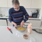 Starejši moški v kuhinji stoji ob pultu in meša rumeno testo v veliki kovinski posodi. Na pultu so še nož, moka in druge sestavine.