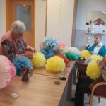 Skupina starejših žensk sedi za mizo v svetlem prostoru, kjer izdelujejo barvite okraske iz svilenega papirja. Okoli njih so razporejene ročno izdelane papirnate pom-pom kroglice v rumenih, modrih, rožnatih in oranžnih odtenkih. Na mizi so tudi barvni svinčniki, vrvice in škarje. Ena od žensk drži v rokah skoraj dokončan okras v rožnato-beli barvi.