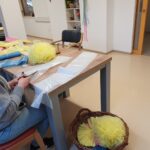 Mlada ženska v sivi jopici in očalih sedi za mizo ter reže liste barvite plastike, ki jih uporablja za izdelavo okraskov. Na mizi so razgrnjeni različni materiali v rumeni, beli in modri barvi. Ob mizi stoji košara, napolnjena z rumenimi papirnatimi pom-pom kroglami. V ozadju, v hodniku, sedi rjavkast maček in gleda proti vratom.
