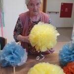 Starejša ženska s sivimi lasmi in roza brezrokavnikom stoji za mizo ter drži v rokah velik rumen papirnat okras. Pred njo so razporejeni še drugi podobni okraski v rumeni, modri in oranžni barvi. Videti je ponosna na svoje delo, medtem ko pozira za fotografijo.