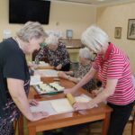 Skupina starejših žensk se zbrano posveča peki. Dve gospe stojita in z valjarjema razvaljujeta testo, medtem ko preostale tri sedijo za mizo in oblikujejo rolice. Na mizi so peki papir in pladenj z že pripravljenimi rolicami iz testa.