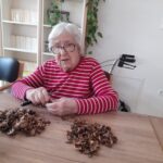 Oseba v rdeči črtasti majici sedi za mizo in ročno trga orehe. Ob njej je večja količina orehovih lupin in majhen nož.