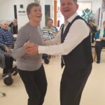 Starejša gospa v sivem puloverju pleše s starejšim moškim v črnem brezrokavniku in beli srajci. Oba se smejita in gledata v kamero. V ozadju so ostali stanovalci doma, nekateri sedijo, nekateri opazujejo dogajanje. Prostor je okrašen z rdečimi papirnatimi cvetovi in visečimi dekoracijami iz vrvic.