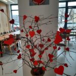 Na sredi prostora stoji visoka vaza iz pletene slame, v kateri so postavljene suhe veje. Na vejah so pritrjeni številni rdeči papirnati srčki različnih velikosti. V ozadju je videti jedilnico z mizami in stoli ter stekleno steno, skozi katero se vidi zunanjost. Na steni nad vejami je pritrjena slika dveh rok, ki držita rdeče srce.