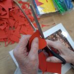 Na mizi ležijo razrezani rdeči papirji. V ospredju starejše roke s škarjami izrezujejo rdeč papirnat srček. Na mizi so tudi drugi ustvarjalni materiali, kot so barvni papirji, škatle in fotografija.