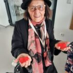 Starejša gospa z očali, črnim klobukom in elegantnim črnim plaščem stoji in se nasmejano ozira v kamero. Okoli vratu nosi rožnato-črn šal, v rokah pa drži dva rdeča papirnata srčka.