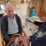 Starejši gospod s sivimi lasmi in toplim nasmehom sedi na stolu, medtem ko mu starejša gospa v temni majici iz košare podaja zavit darilni paket s klobaso in suhim sadjem. Dogodek se odvija v svetlem prostoru, v ozadju so mizice in naslanjači.