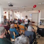 Skupina starejših oseb sedi v krogu v svetlem prostoru, pozorno poslušajo mlajšo žensko v beli majici, ki jim nekaj razlaga. Nekateri stanovalci sedijo na stolih, drugi v invalidskih vozičkih. Prostor je lepo okrašen z rdečimi papirnatimi cvetovi, na stenah so knjižne police in dekoracije.