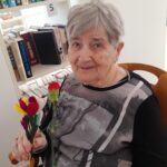 Starejša gospa s kratkimi sivimi lasmi in nasmehom na obrazu sedi pred knjižno polico ter v rokah drži šopek pisanih rož. Oblečena je v črno-sivo majico s sodobnim vzorcem. Knjižna polica v ozadju je napolnjena z različnimi knjigami.