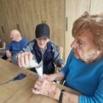 Starejši moški s črno kapo previdno preliva riž skozi papirnati lijak v blago vrečko, ki jo drži starejša gospa v modri majici. Oba sta osredotočena na nalogo. V ozadju sedita še dva starejša moška in opazujeta dogajanje.