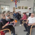 Vadba z blazinicami za motoriko poteka v večjem prostoru z belimi stenami, slikami na steni in pisanimi okraski na stropu. Starejši udeleženci, nekateri v invalidskih vozičkih, si izmenjujejo mehke blazinice različnih barv in oblik.