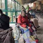 Skupina starejših oseb, večina v invalidskih vozičkih, sedi na pokriti terasi doma za starejše. So toplo oblečeni, nekateri z odejami, pred njimi so barvni plastični kozarci. V ozadju nekaj zaposlenih.