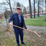 Starejši gospod v trenirki in s kapo drži grablje, stoji ob ograji ob gredici. Pogled ima resen, v ozadju je potka in drevesa brez listja.