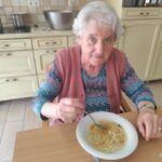 Starejša ženska s sivimi lasmi sedi za leseno mizo in je krožnik domače juhe z rezanci. V ozadju je kuhinja z rumenimi omaricami in lonci na štedilniku. Pogled ji je usmerjen proti kameri, na obrazu ima blag nasmeh.